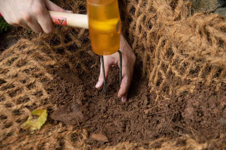 Using weed mat pins with a hammer to secure natural fiber matting into soil for gardening