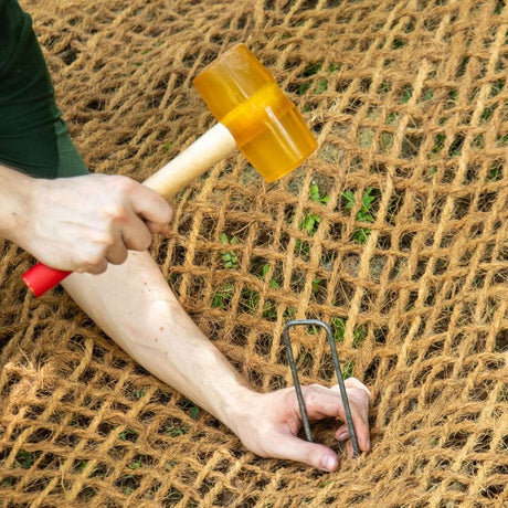 Hand using a yellow mallet to secure weed mat pins into a natural fiber landscaping mat