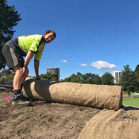 Worker rolling out HEIGER jute matting on a grassy outdoor area under blue sky